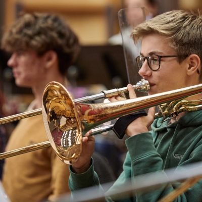 A boy with blonde hair and wearing a green jumper is sitting down playing a trombone